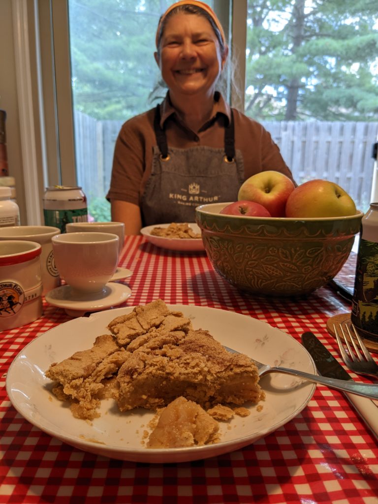 A slice of apple pie on a plate in the foreground, with Jackie smiling in the background.