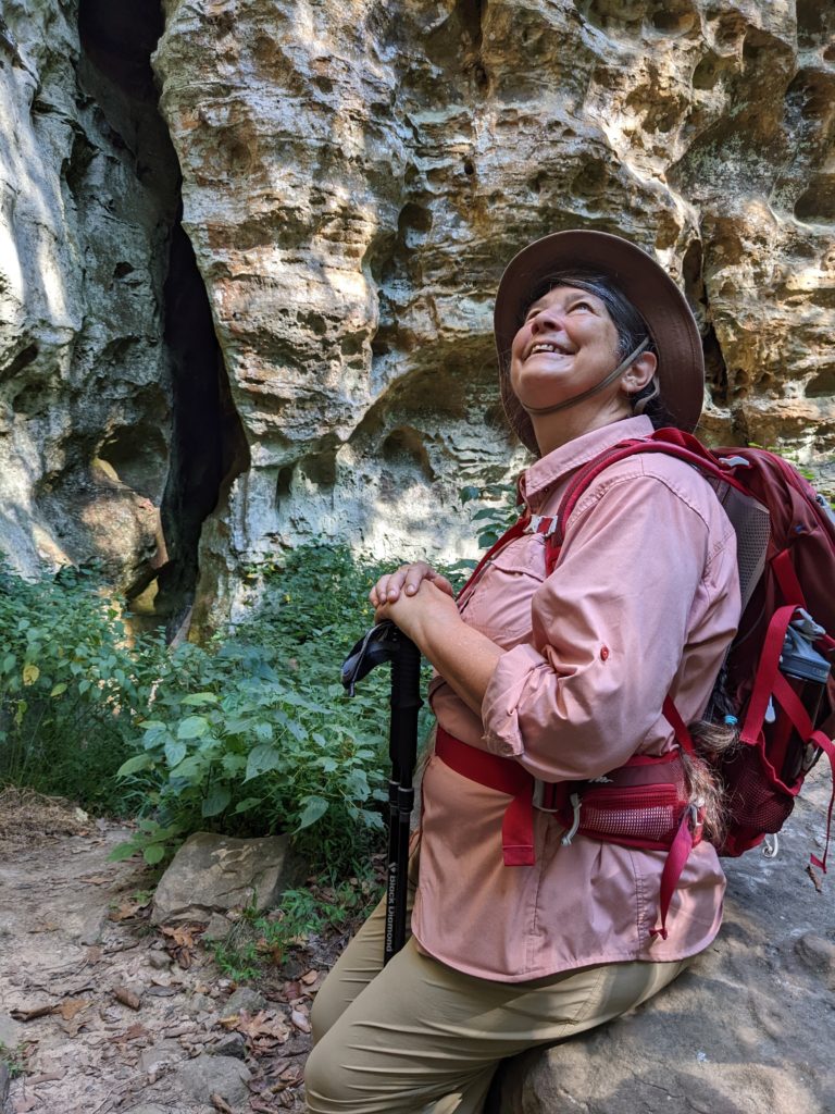 Jackie in a backpack with her trekking poles sitting on a rock on the Giant City Trillium Trail, looking up at rock walls