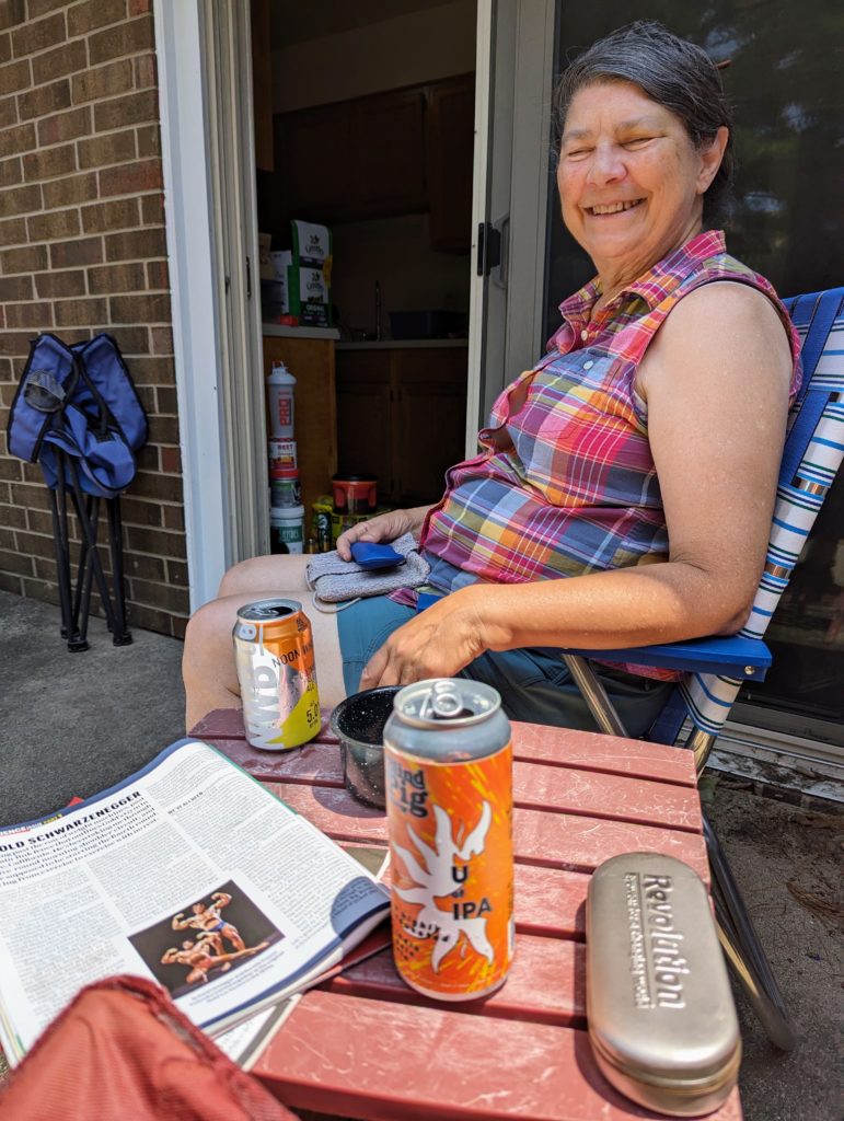 Jackie (with her eyes closed) beyond a small table with magazines, beers, and a sunglasses case