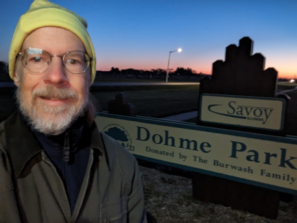 Selfie wearing a high-viz hat, standing in front of the Dohme Park sign at dawn