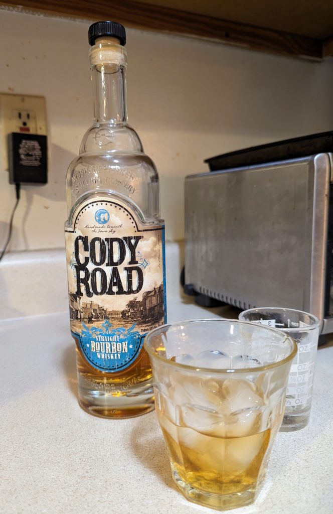 A bottle of Cody Road bourbon behind a glass of the whiskey on the rocks.