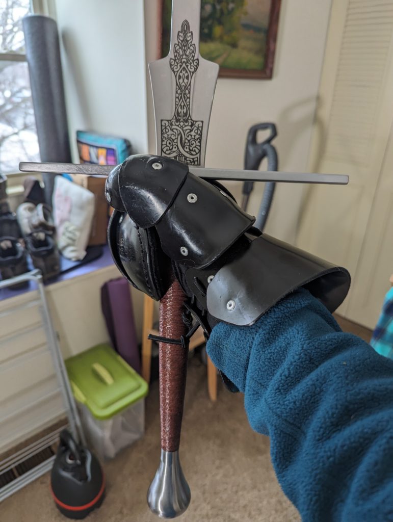 The hilt and schilt of a longsword, held by one hand in a heavy glove, showing the fancy etching on the schilt