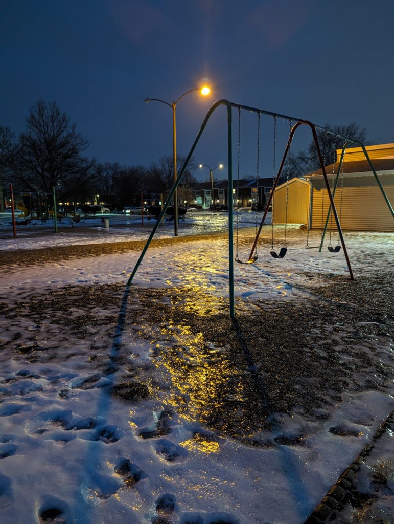 Street lights shine on snow and ice, with a swing set in the foreground.