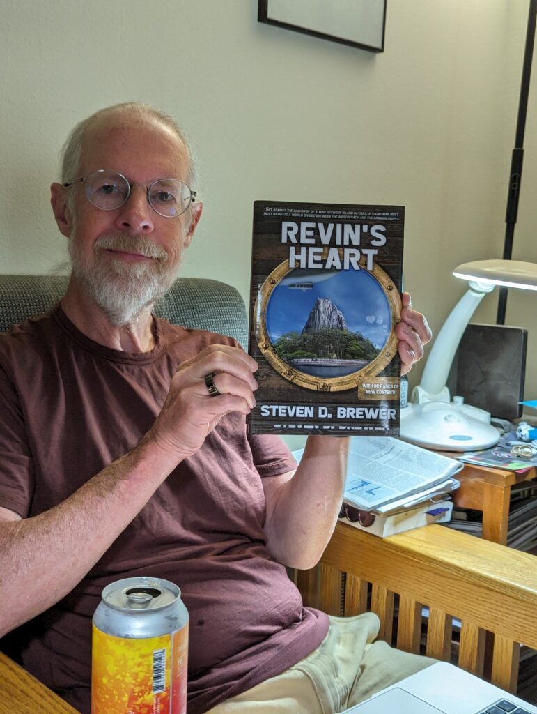 Me sitting in my chair behind my laptop computer and a beer, holding a hardback copy of Revin's Heart by Steven D. Brewer