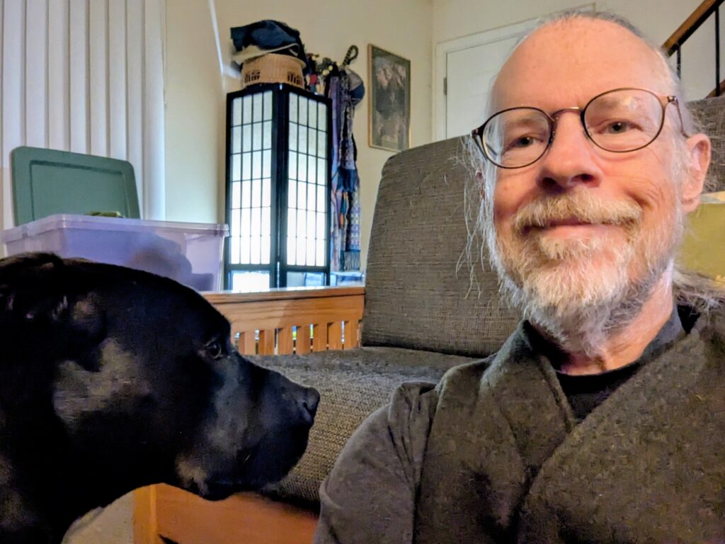 Me sitting on the floor wearing a wool vest, with the head of a black dog facing me