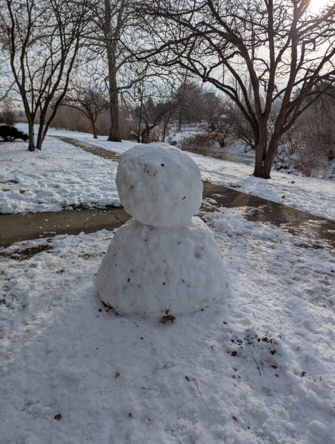 A rather blank-faced snowman, standing next to the sidewalk, with snow and some trees beyond, and a creek beyond that.