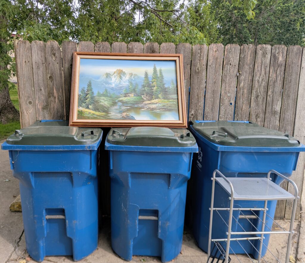 A landscape painting in a frame balaned on top of the recycling bins near the dumpster